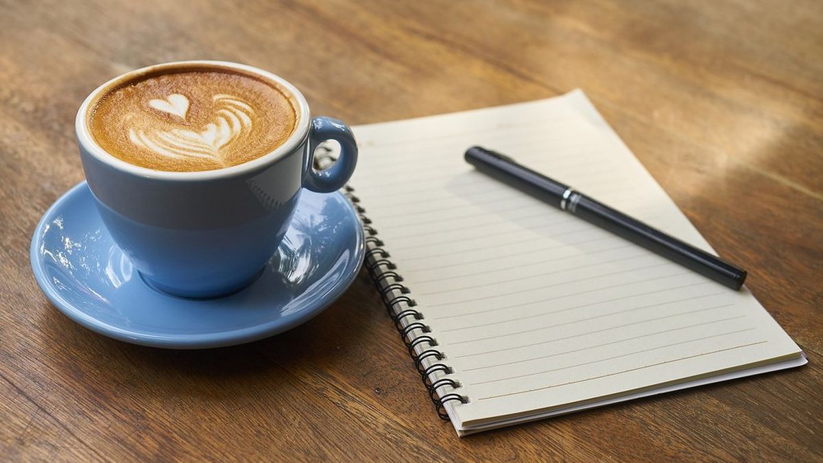 Cup of coffee beside an open lined notebook and pen on a wooden table